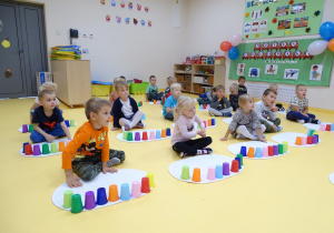 Dzieci siedzą przed białymi dywanikami i układają kolorowe kubeczki